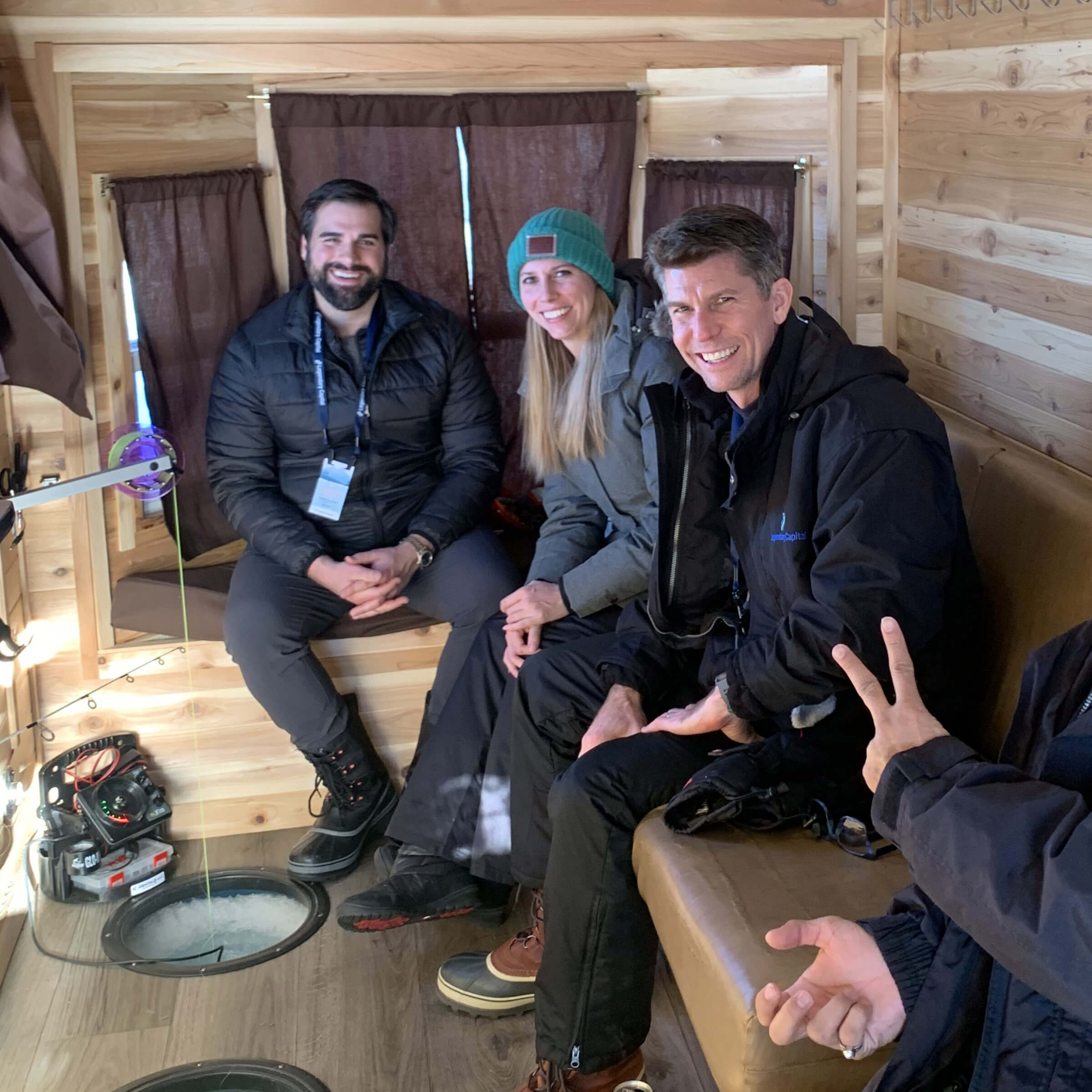 A group photo of three smiling people in winter clothes sitting in a fish shack.