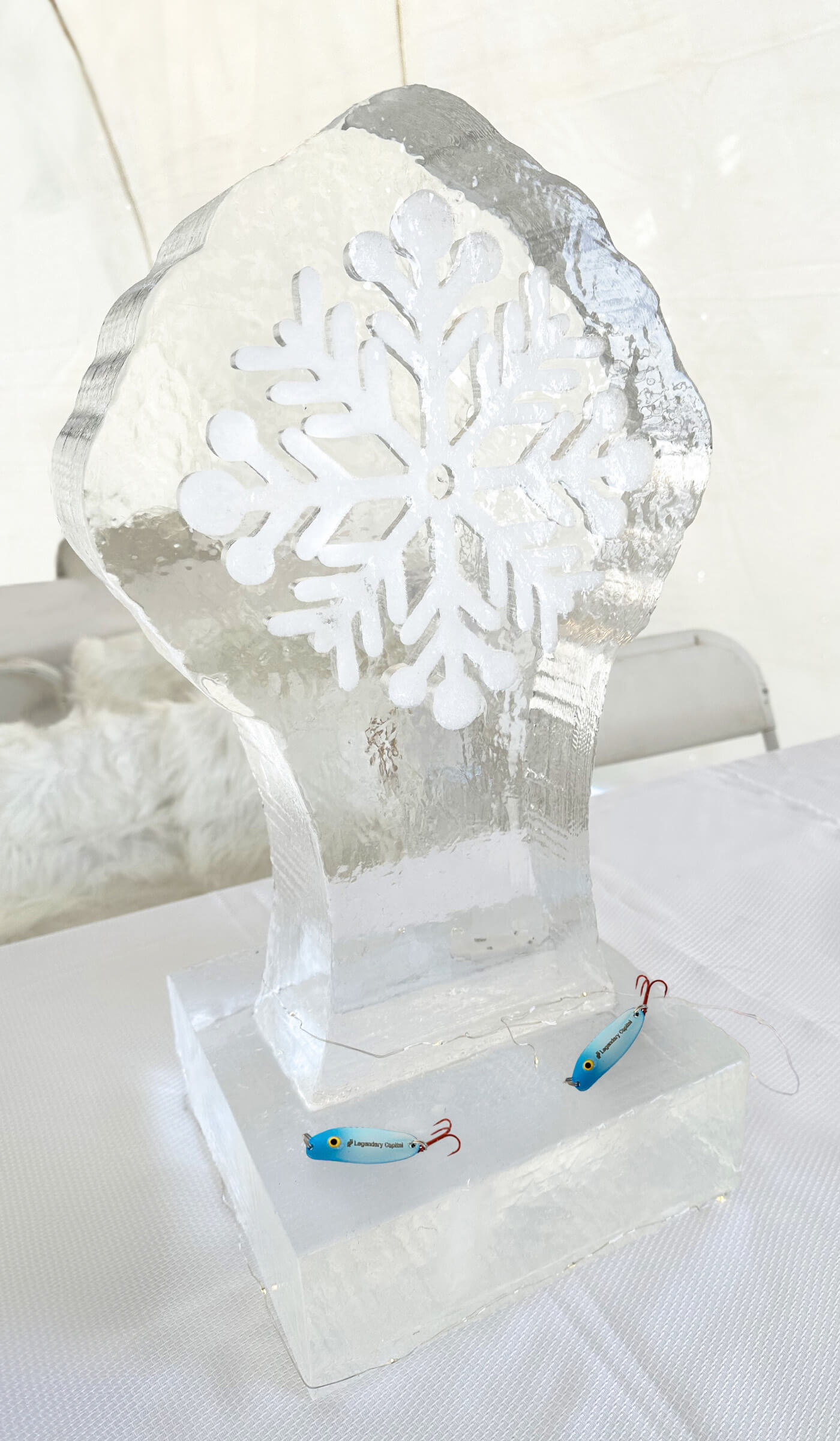 An ice sculpture with a large, white snowflake in the center and two blue fishing lures on the base.