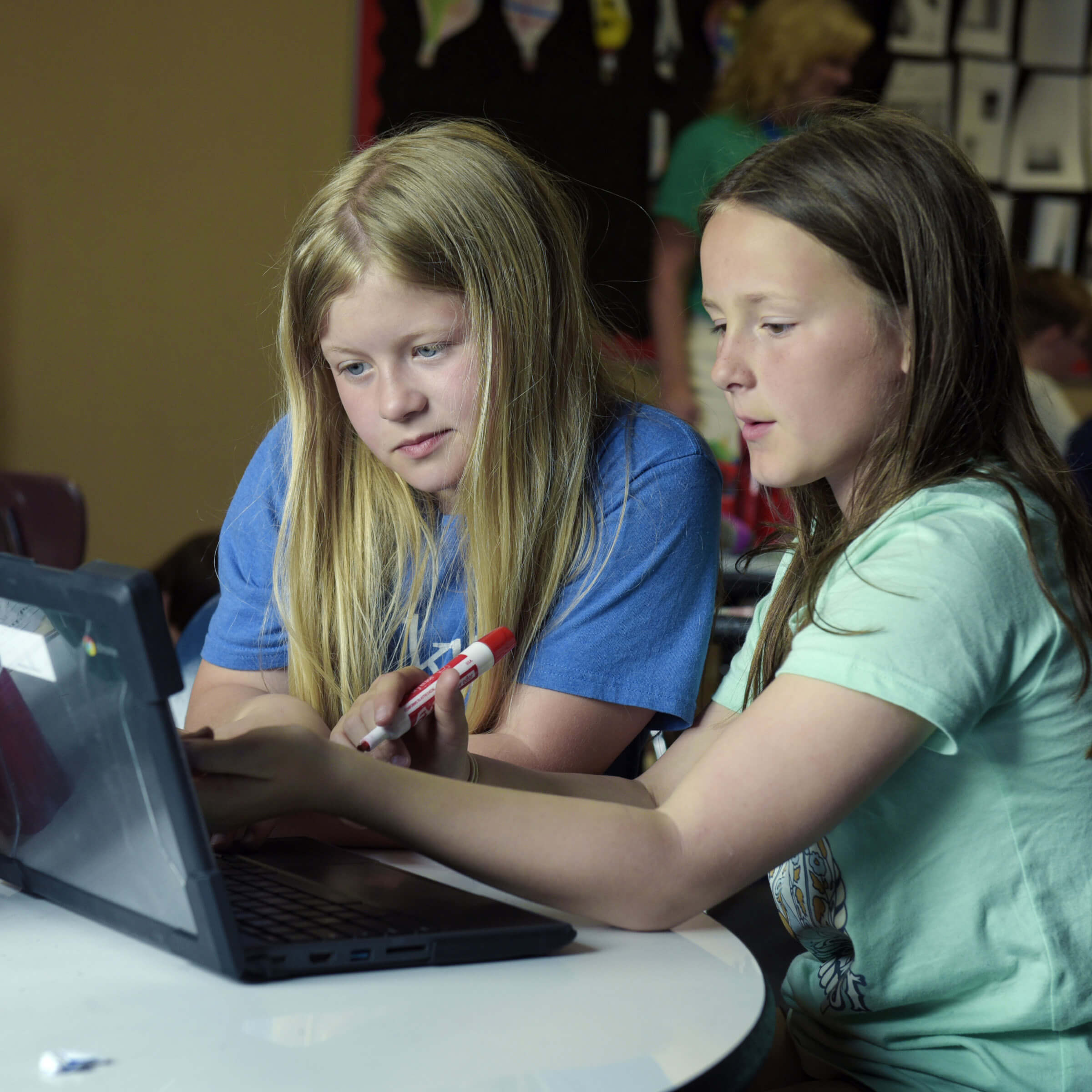 A group of two girls sitting at a school table looking at a laptop.
