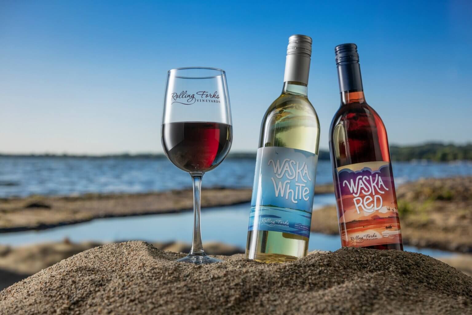 White and red wine bottles with a wine glass in the sand in front of lake